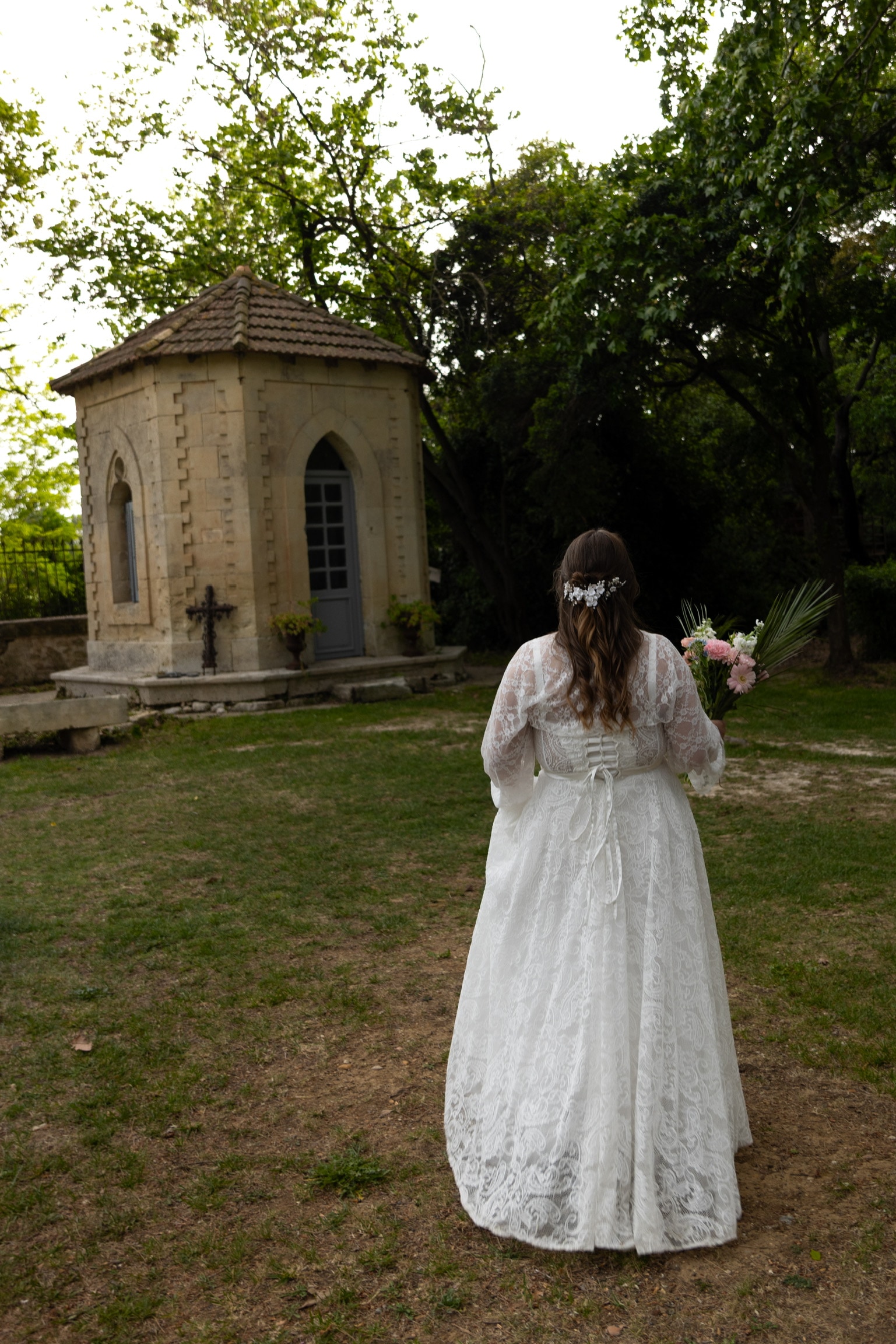 La mariée, de dos, marche vers une petite chapelle en pierre, bouquet à la main
