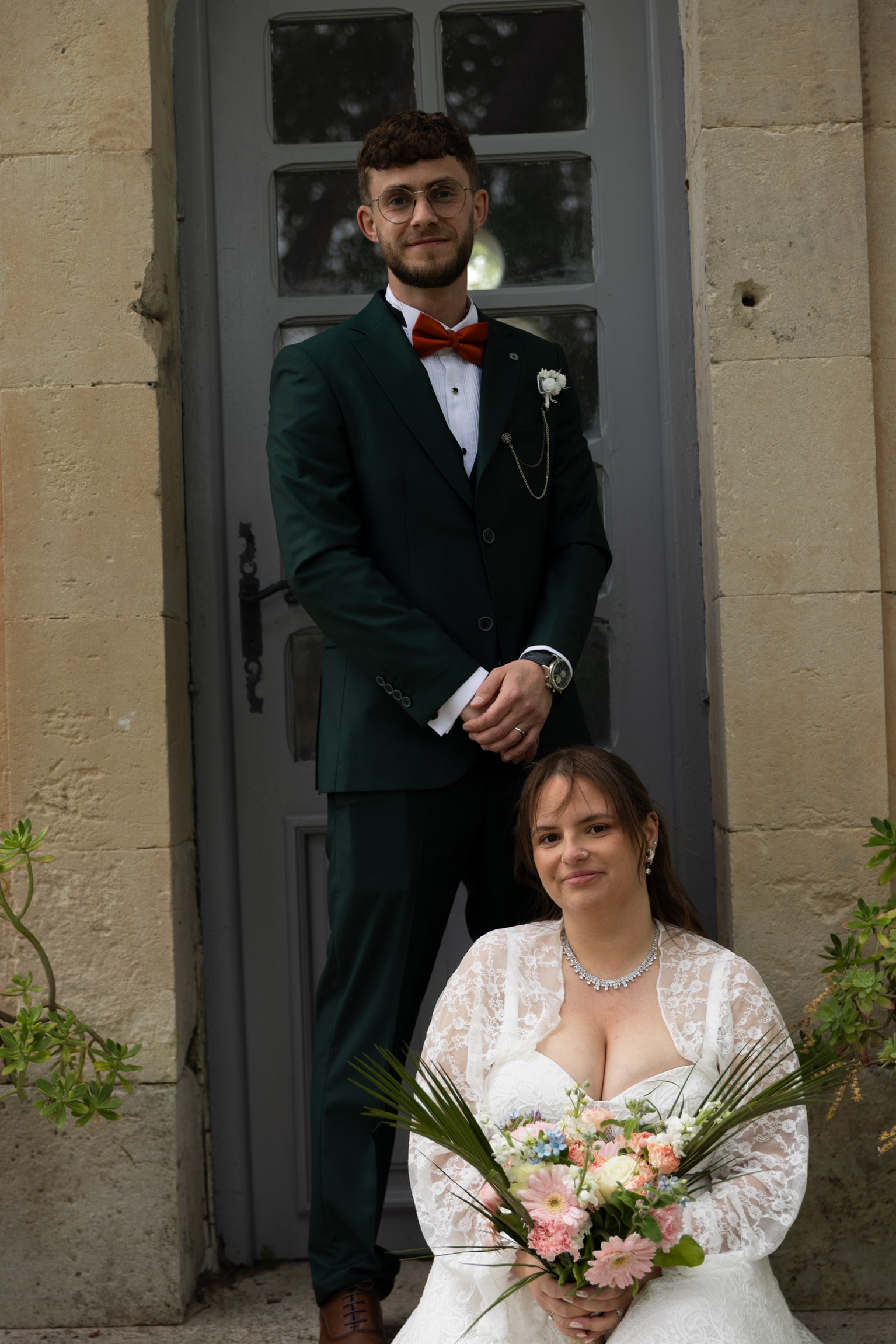 Le marié debout derrière la mariée assise devant une porte grise